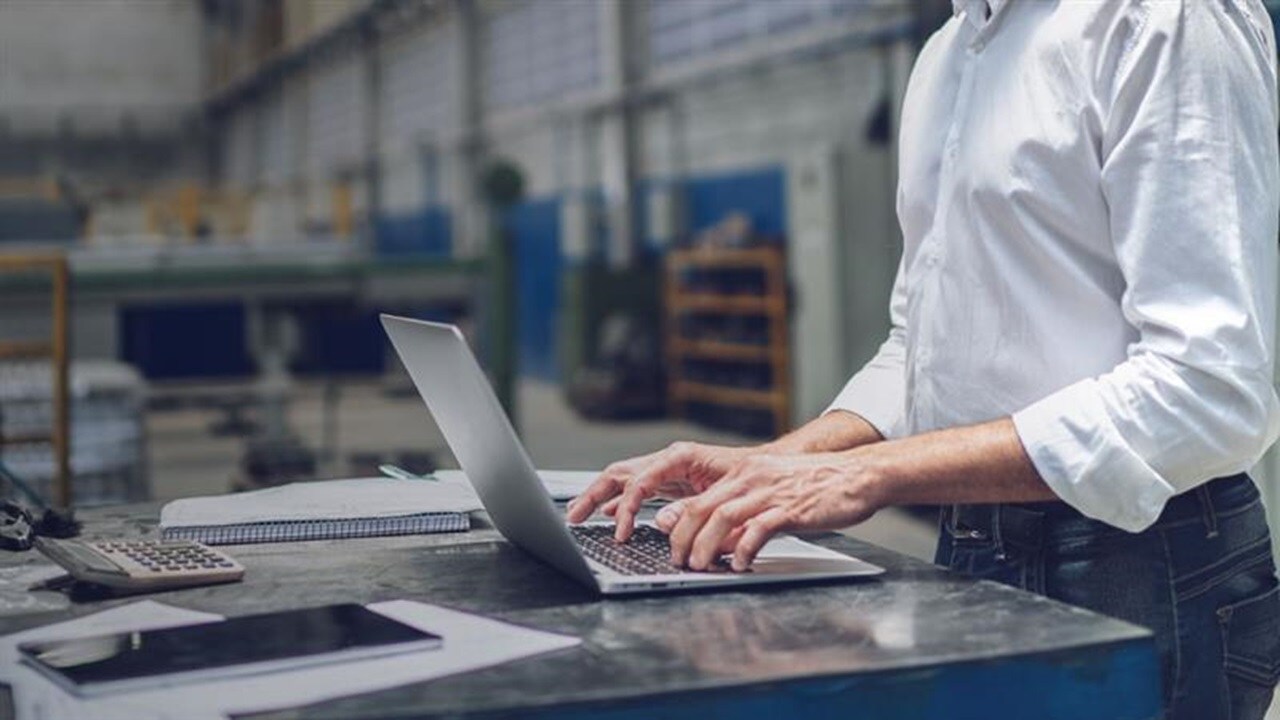 A person in a white shirt using a laptop on a workbench in an industrial warehouse, with a notebook, calculator, and tablet nearby.