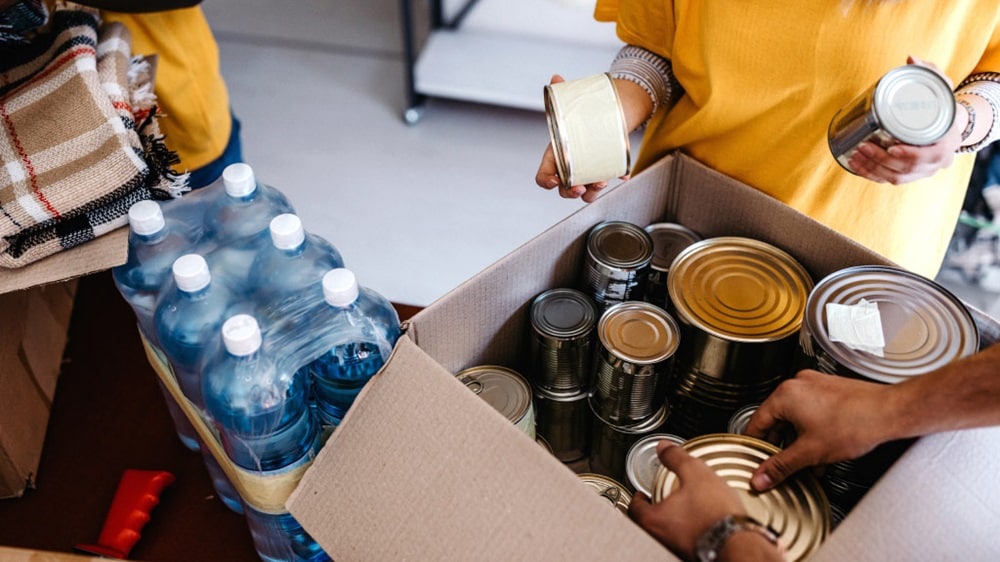 Volunteers sort canned food and bottled water in a cardboard box during a community support event.