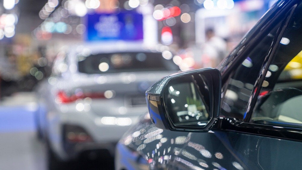Close-up of a car's side mirror, with a blurred silver car in the background at a vibrant auto show.
