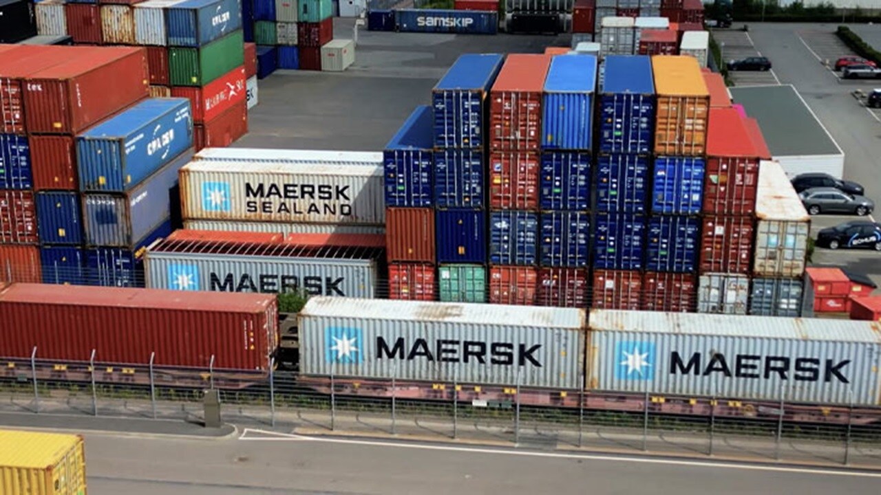 Maersk containers on a single railcar passing a stacked container yard, with various colored shipping containers in the background.