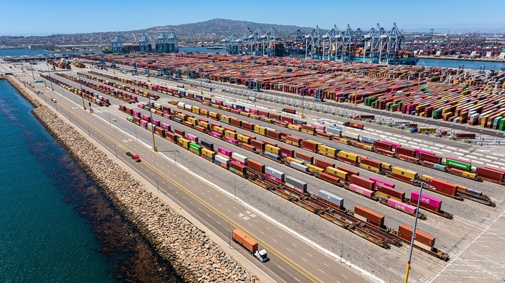 Aerial view of a large container terminal with stacked shipping containers, cranes, rail lines and trucks along the waterfront.