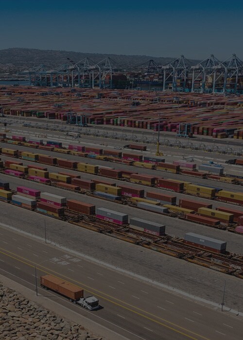 Aerial view of a large container terminal with stacked shipping containers, cranes, rail lines and trucks along the waterfront.