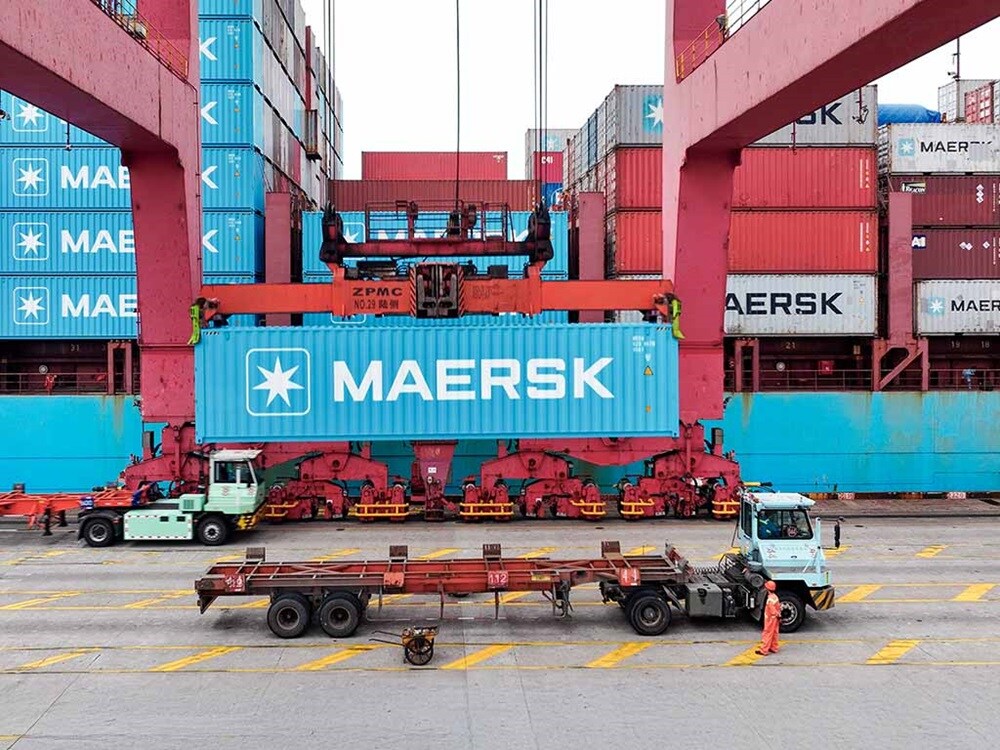 Contenedor de transporte de Maersk levantado por una grúa pórtico en una terminal de puerto de orígenes con camiones debajo